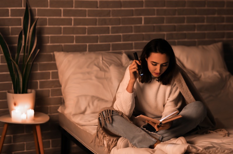 young woman reading a book with flashlight in bedroom during power outage